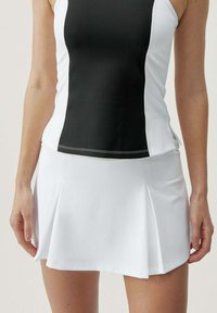 Mujer vistiendo una camiseta deportiva sin mangas en blanco y negro y una falda de tenis blanca con pliegues, de pie con los brazos relajados a los lados.