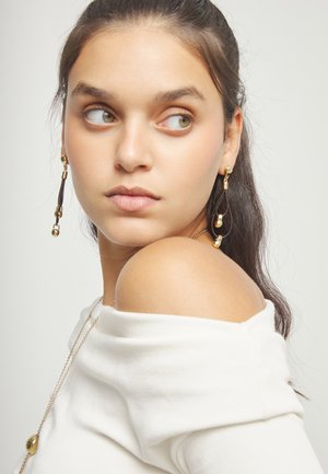Joven con el cabello oscuro recogido mirando hacia un lado, vestida con una blusa blanca fuera de los hombros, largos pendientes dorados y un collar dorado.