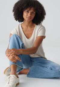 Camicia beige a maniche corte con scollatura ampia, abbinata a jeans a gamba larga blu chiaro e sneakers bianche con suola in gomma, in posizione seduta.