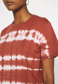 T-shirt couleur rouille avec un motif tie-dye en blanc, manches courtes, col rond et texture en coton douce.