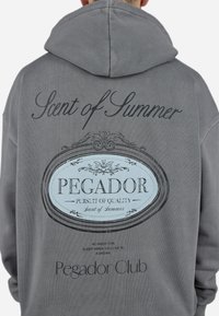 Sudadera gris con un gráfico central que incluye el texto: "Scent of Summer" y "PEGADOR," adornada con detalles ornamentales. Textura suave.