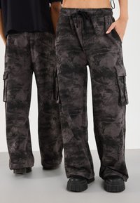 Two people wearing black and gray camo cargo pants and black platform shoes standing side by side on a gray floor.