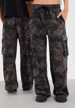 Due persone indossano pantaloni cargo a carico mimetico neri e grigi e scarpe con plateau nere, in piedi affiancate su un pavimento grigio.
