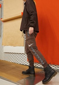 Brun vindjacka med dragsko, bruna joggers med vita sidostråk och cargo-fickor, samt svarta snörstövlar med tjock sula.