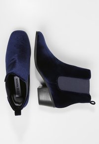 Botines de terciopelo azul marino con tacón cuadrado, panel lateral elástico y una pestaña en la parte posterior para ponérselos con facilidad. Textura suave.