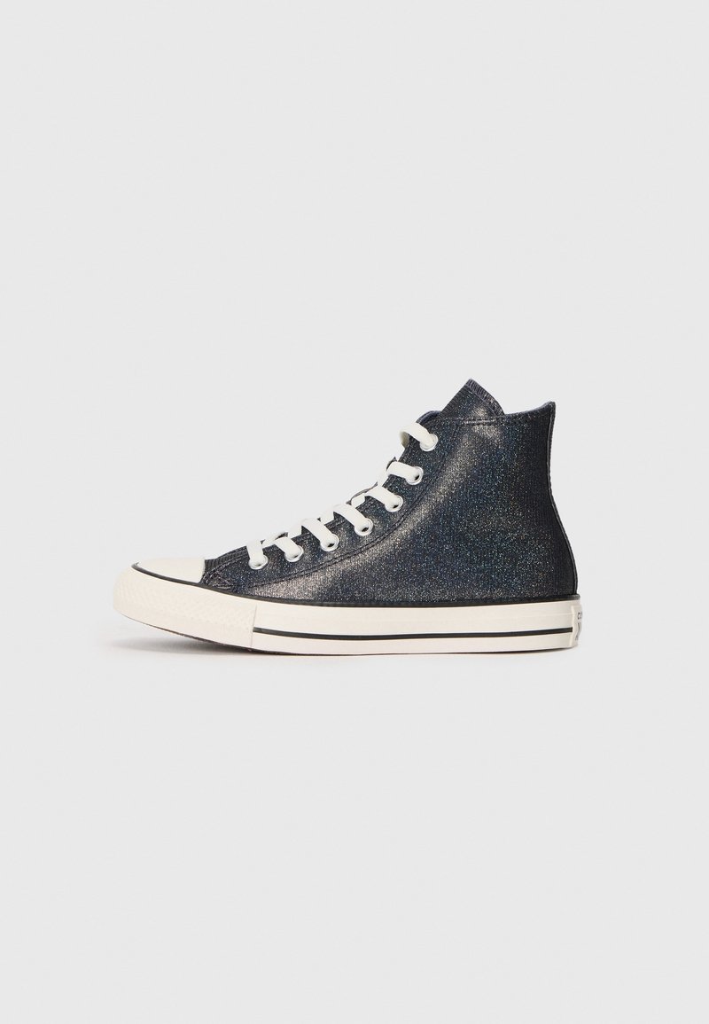 Baskets montantes noires avec semelle et bout en caoutchouc blanc, lacets blancs et texture pailletée subtile sur fond gris clair.
