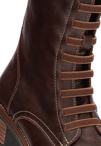 Botín de cuero marrón con una textura suave, que presenta cordones marrones y costuras en contraste a lo largo de los bordes y ojales.