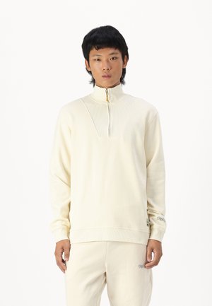 Cream-colored fleece sweatshirt with a high collar, half-zip, ribbed texture on upper section, and logo detail on the sleeve.