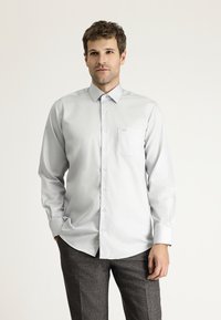Camicia button-up a maniche lunghe grigia chiara con colletto strutturato, taschino sul petto e texture del tessuto liscia, abbinata a pantaloni grigi con fantasia.