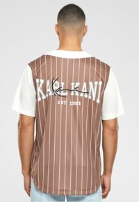 Brun stribet trøje med korte hvide ærmer. Har "KARL KANI" og "EST. 1989" trykt på ryggen i hvid og sort skrift.