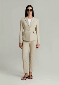 Blazer beige su misura e pantaloni abbinati, realizzati in un tessuto morbido, dotati di tasche e chiusura con un singolo bottone. Indossato con una t-shirt bianca e sandali.