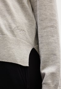 Pull en maille beige avec ourlet côtelé et petite fente latérale, logo brodé près de la taille, porté sur un pantalon noir.