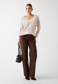 Beżowy sweter z dekoltem w serek o luźnym kroju, brązowe jeansy z wysokim stanem oraz brązowe botki na obcasie. Dodatki to torebka we wzory i minimalistyczna biżuteria.