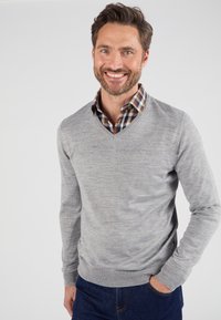 Maglione grigio chiaro a collo a V realizzato in materiale knit, con un design aderente. Indossato sopra una camicia a quadri, con polsini e vita visibili.