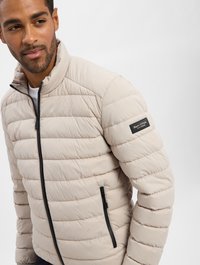 Beige gewatteerde jas met horizontaal gestikt design, zwarte ritszakken, staande kraag en logo patch op de linker mouw.