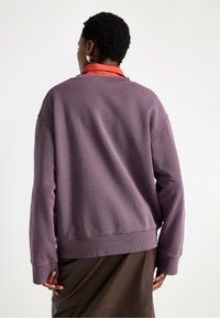 Felpa viola con texture a coste, vestibilità rilassata e maniche lunghe. Indossata sopra una gonna marrone, presenta un collo rotondo e spalle scese.