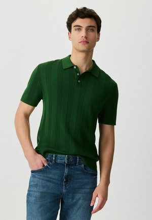 Junger Mann mit kurzen lockigen Haaren, der ein grünes, kurzärmliges Strick-Poloshirt und blaue Jeans trägt, steht mit einer Hand in der Tasche.