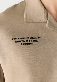 Beige polo shirt with a collar, featuring a black printed text: "LOS ANGELES COUNTY SANTA MONICA SOUNDS" on the front. Soft texture.