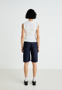 White textured top with frill sleeves and ruched details at the back, paired with dark blue denim bermuda shorts. Black shoes and light socks.