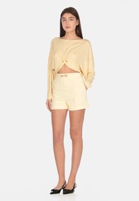Top corto giallo chiaro con dettaglio annodato, abbinato a pantaloni corti a vita alta coordinati con una fibbia in tonalità oro. Scarpe slingback nere.