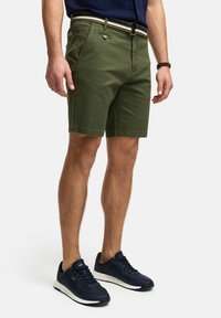Olijfgroene katoenen shorts met een klassieke pasvorm, voorzien van een sluiting met een voorste knoop, riemlussen en een gestreepte stoffen riem. Gecombineerd met marineblauwe sneakers.