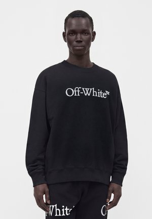 Νέος μαύρος άνδρας με μαύρο φούτερ Off-White και αντίστοιχο παντελόνι με λευκό λογότυπο, στέκεται σε απλό λευκό φόντο.