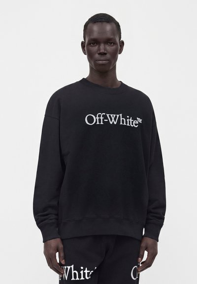 Νέος μαύρος άνδρας με μαύρο φούτερ Off-White και αντίστοιχο παντελόνι με λευκό λογότυπο, στέκεται σε απλό λευκό φόντο.