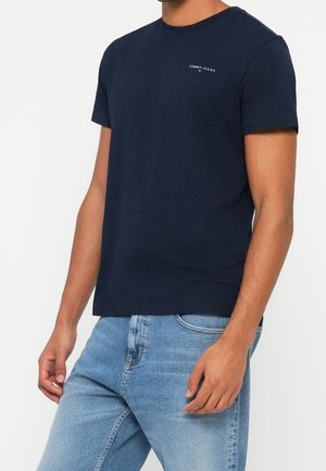 Mężczyzna w granatowym krótkim rękawie koszulki Tommy Jeans i jasnoniebieskich dżinsach, stojący na prostym białym tle.