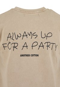Beige bomulls-T-skjorte med svart tekst som sier "ALWAYS UP FOR A PARTY" og "ANOTHER COTTON" trykt på baksiden. Enkel rund hals-design.