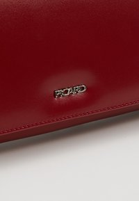 Article en cuir rouge avec des coutures soignées et un petit logo en métal argenté portant l'inscription "PICARD" sur une surface blanche.