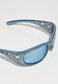 Saulesbrilles ar zilu plastmasas rāmi, izliekta dizaina un ziliem objektīviem. Rāmī ir ventilācijas caurumi un kontrastējoši melni akcenti.