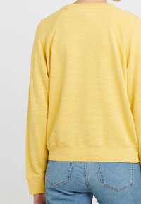 Felpa gialla con scollatura a girocollo a coste, maniche raglan e tessuto testurizzato. Abbinata a jeans blu, che mostrano la vista da dietro.