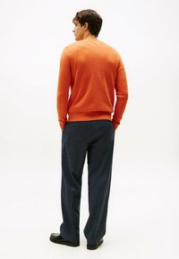 Oranges Wollpullover mit klassischer Passform, V-Ausschnitt und gerippten Bündchen, kombiniert mit dunkelblauen weit geschnittenen Hosen und schwarzen Schuhen.