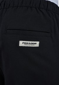 Pantalons noirs avec une taille élastique et une poche arrière arborant une étiquette blanche sur laquelle est inscrit "PEGADOR" en texte noir. Le matériau semble lisse et durable.