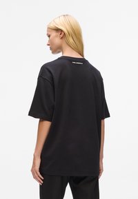 Femme aux longs cheveux blonds portant un t-shirt noir ample de Karl Lagerfeld, vue de dos sur un fond blanc uni.