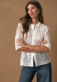 Balta mežģīņu blūze ar ziedu rakstiem, garām piedurknēm ar uzlocītiem manžetēm un pogām priekšpusē. Valdziņota kopa ar zilām džinsa biksēm.