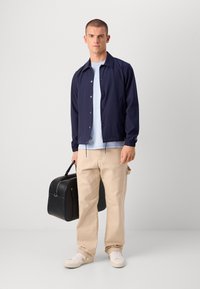 Navy windbreaker with a collar, button closure, and elastic cuffs; light blue t-shirt underneath; beige pants; black bag; white sneakers.
