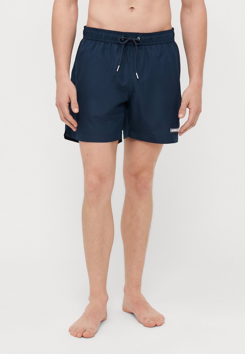 Shorts de bain bleu marine avec une taille élastique, cordon de serrage et un patch logo sur le côté. Tissu lisse et léger, longueur mi-cuisse.