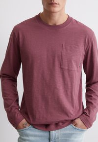 Langärmliges, burgunderfarbenes Hemd aus weichem, strukturiertem Stoff mit einer Brusttasche und geripptem Rundhalsauschnitt, kombiniert mit hellblauen Jeans.