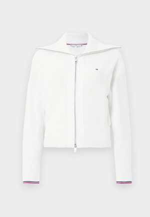 Biała dzianinowa kardigan z zamkiem błyskawicznym i szerokim kołnierzem, o prążkowanej fakturze, z kontrastowymi czerwonymi i niebieskimi paskami na mankietach. Posiada srebrny zamek.