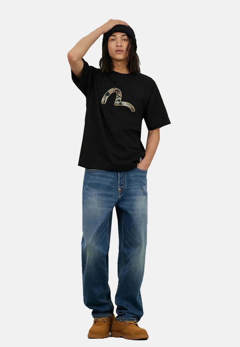 Sort t-shirt med et grafisk logo, parret med lyseblå denimjeans og brune støvler. Modellen har langt, tekstureret hår og en sort hue.