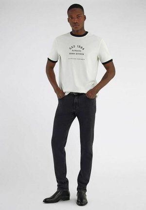 Giovane uomo con t-shirt bianca con grafica e bordo nero, jeans slim neri e stivali in pelle neri, in piedi con le mani in tasca.