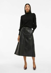 Femme debout en pull à col roulé noir, jupe midi en cuir noir et escarpins à bout pointu marron sur fond blanc.