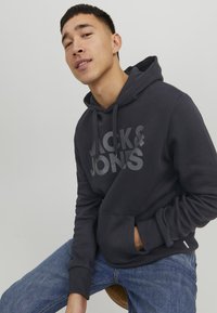 Felpa grigia scura con grande logo "JACK & JONES". Presenta una tasca a marsupio, cappuccio con cordino e polsini a coste; realizzata in morbido materiale tessile.