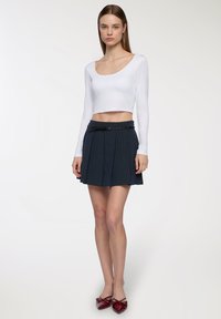 Biała, długa bluzka crop top z długim rękawem w połączeniu z plisowaną granatową spódnicą z paskiem. Modelka nosi czerwone czółenka na wysokim obcasie z kokardką.