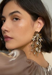 Gold floral earrings with crystals, featuring cascading designs and a mix of leaves and flowers, worn by a person with shoulder-length hair.