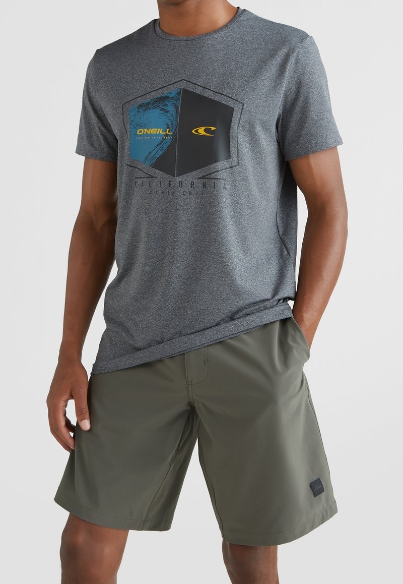 Homme portant un t-shirt graphique gris O'Neill et un short vert olive, les mains dans les poches, sur un fond uni.