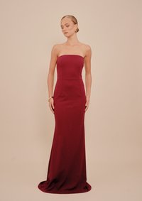 Γυναίκα φορώντας μία strapless, μακριά μπορντό φορεσιά, με χρυσά σκουλαρίκια και βραχιόλι, που στέκει μπροστά σε ένα μπεζ φόντο.