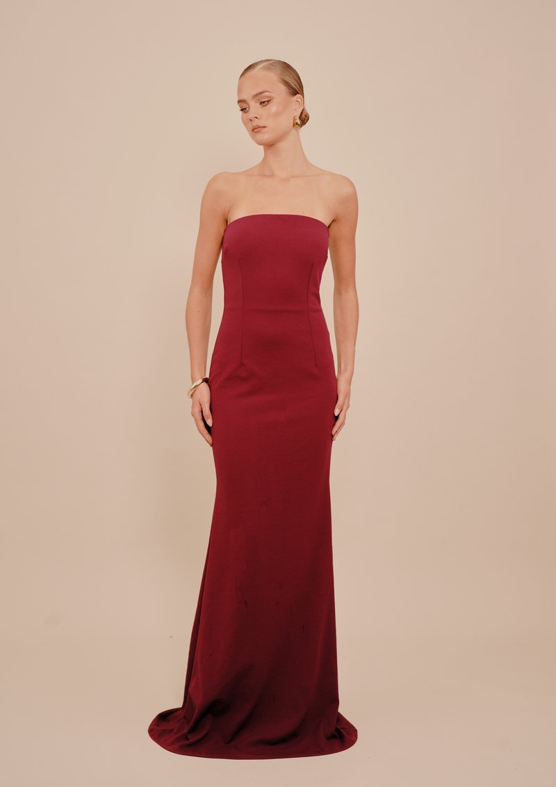 Γυναίκα φορώντας μία strapless, μακριά μπορντό φορεσιά, με χρυσά σκουλαρίκια και βραχιόλι, που στέκει μπροστά σε ένα μπεζ φόντο.