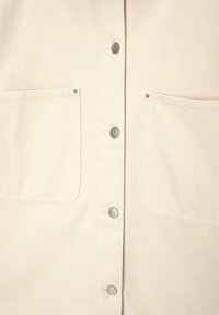 Beige knoopshirt van katoen, met twee voorzakken, zilveren knopen en een textiele stof met zichtbare stiksels.